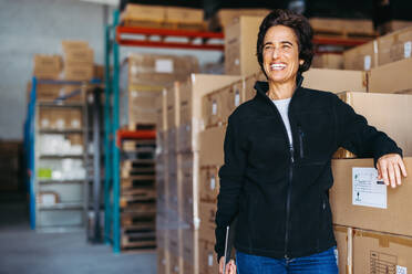 Happy logistics manager looking away with a smile while standing next to a stack of cardboard boxes. Cheerful mature woman working in a large distribution warehouse. - JLPPF01316