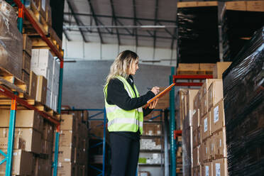 Female warehouse worker writing on a clipboard while standing in front of a stack of cardboard boxes. Woman in a reflective jacket taking inventory in a large distribution centre. - JLPPF01320