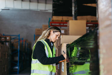 Warehouse supervisor writing on a clipboard while standing in front of a stack of package boxes. Woman in a reflective jacket taking stock in a large distribution centre. - JLPPF01321