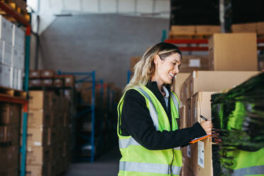 Happy warehouse manager smiling while writing on a clipboard in a large distribution centre. Cheerful young woman doing stock control in a modern fulfillment centre. - JLPPF01322