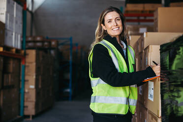 Female warehouse worker smiling at the camera while writing on a clipboard in a distribution centre. Cheerful young woman doing inventory control in a large fulfillment centre. - JLPPF01323