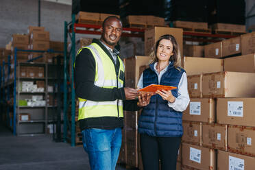 Logistics employees smiling at the camera while holding a bill of lading. Happy freight forwarder receiving goods for shipment from a consignor. Female warehouse manager importing merchandise. - JLPPF01325