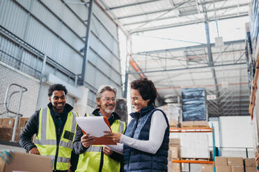 Fröhlich lächelnde Lagerarbeiter, die sich während einer Personalversammlung einen Bericht ansehen. Eine Gruppe verschiedener Mitarbeiter, die gemeinsam ein erfolgreiches Fulfillment-Center betreiben. - JLPPF01329