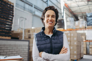 Portrait of a female warehouse employee smiling at the camera while standing in a distribution centre with her arms crossed. Mature woman working as a supervisor in a large logistics centre. - JLPPF01335