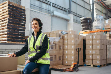 Female warehouse employee looking away with a happy smile while sitting on a rack of cardboard boxes. Cheerful mature woman working as a manager in a large distribution centre. - JLPPF01336