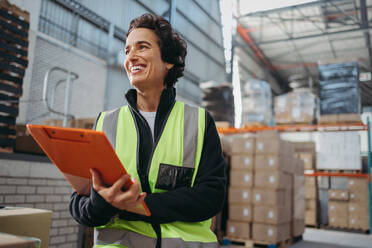 Glücklicher Logistikmanager, der mit einem glücklichen Lächeln wegschaut, während er ein Klemmbrett hält. Reife Frau, die in einem großen Vertriebslager arbeitet. - JLPPF01337