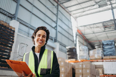 Älterer Logistikmanager lächelt in die Kamera, während er ein Klemmbrett in einem Auslieferungslager hält. Fröhliche Frau, die in einem großen Fulfillment-Center arbeitet. - JLPPF01338