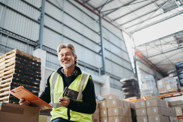 Happy senior man smiling at the camera while holding a clipboard in a distribution centre. Cheerful warehouse manger sitting on a rack of cardboard boxes in a reflective jacket. - JLPPF01339