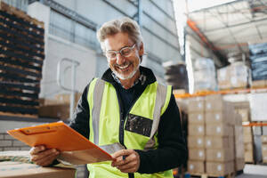 Fröhlich lächelnder Lagerleiter beim Lesen einer Akte in einem Logistikzentrum. Fröhlicher älterer Mann mit reflektierender Jacke in einem großen Vertriebszentrum. - JLPPF01341
