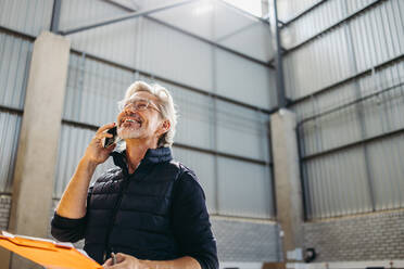 Fröhlicher älterer Mann, der lächelt, während er einen Anruf in einem Auslieferungslager entgegennimmt. Fröhlicher Logistikmanager, der am Telefon in einem großen Erfüllungszentrum Versandpläne erstellt. - JLPPF01343