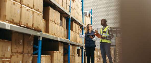 Eine Lagerverwalterin spricht mit ihrem Kollegen, während sie ein digitales Tablet in der Hand hält. Zwei Logistikmitarbeiter erfassen das Inventar mithilfe eines digitalen Lagerverwaltungssystems. - JLPPF01349