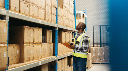 Male warehouse worker recording SKU's on a clipboard while holding a barcode scanner. Logistics worker doing inventory control using a barcode management system in a distribution centre. - JLPPF01352
