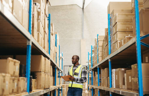 Warehouse worker writing down package details while standing in front of a storage shelf in a fulfilment centre. Young man taking inventory in a large distribution storehouse. - JLPPF01353