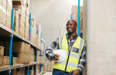 Warehouse order picker smiling happily with a headset around his neck. Cheerful warehouse employee working with voice-picking technology in a modern distribution centre. - JLPPF01366