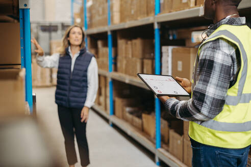 Logistikmitarbeiter verwenden ein Lagerverwaltungssystem auf einem digitalen Tablet. Zwei Lagerarbeiter arbeiten mit einem statistischen Bericht auf ihrem Dashboard für die Bestandsverwaltung. - JLPPF01371