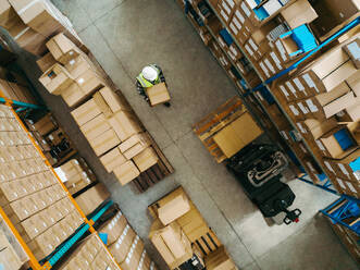 Aerial view of a warehouse worker carrying a package box in a large logistics centre. Top view of a young man moving goods with a pallet jack within a modern fulfillment centre. - JLPPF01372