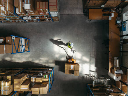 Aerial view of a warehouse employee pulling a pallet truck loaded with cardboard boxes. High angle view of a man in a reflective jacket taking goods for storage in a logistics centre. - JLPPF01373