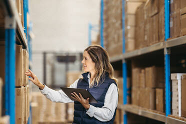 Cheerful young woman checking shipping labels against an inventory list on a digital tablet. Happy logistics worker doing stock control using smart warehousing technology. - JLPPF01387