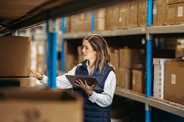 Female warehouse worker checking shipping labels against an inventory list on a digital tablet. Woman doing stock control using warehouse management software in a distribution centre. - JLPPF01388