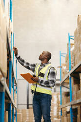 Male logistics worker checking the labels of package boxes stored at the top of a warehouse shelf. Young black man doing inventory control in a large distribution centre. - JLPPF01393