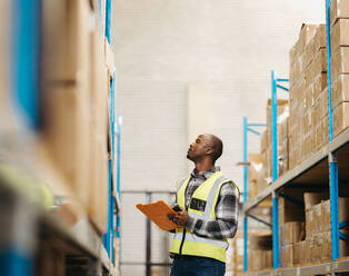 Junger Logistikmitarbeiter, der in einem großen Lagerhaus die Details von Paketboxen oben auf einem Regal überprüft. Junger Mann bei der Bestandskontrolle in einem modernen Vertriebszentrum. - JLPPF01394