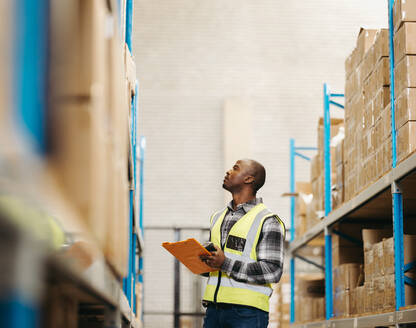Young logistics worker checking the details of package boxes at the top of a storage shelf in a large warehouse. Young man doing inventory control in a modern distribution centre. - JLPPF01394