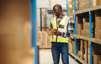 Happy warehouse manager writing on a clipboard while standing next to a storage shelf in a logistics centre. Smiling man doing inventory control in a modern distribution storehouse. - JLPPF01395