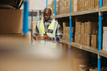 Black logistics manager writing on a clipboard while standing next to a storage shelf in a modern warehouse. Young man doing inventory control in a large distribution centre. - JLPPF01396