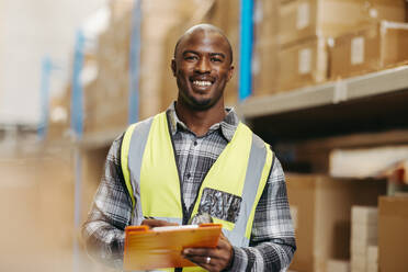 Cheerful young warehouse manager smiling at the camera while standing in between storage shelves in a logistics centre. Happy young man taking inventory in a large distribution storehouse. - JLPPF01398