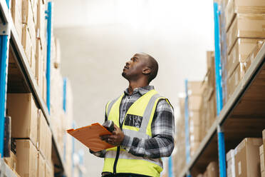 Young logistics supervisor checking the details of package boxes stored at the top of a warehouse shelf. Young man doing stock control in a modern distribution centre. - JLPPF01399