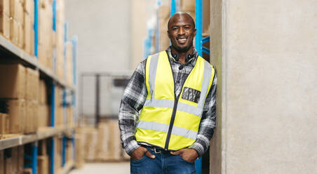 Portrait of a warehouse worker smiling at the camera with a headset around his neck. Happy logistics employee working with voice-picking technology in a large fulfillment centre. - JLPPF01403