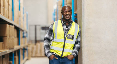 Cheerful warehouse voice picker smiling at the camera with a headset around his neck. Happy logistics worker working with voice-picking technology in a modern distribution centre. - JLPPF01404