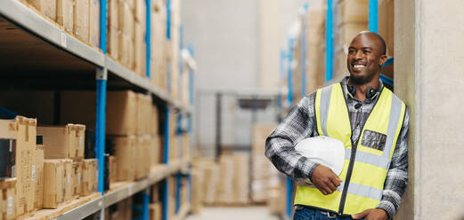 Fröhlicher Lagerkommissionierer, der mit einem Headset um den Hals lächelnd in die Ferne schaut. Fröhlicher Logistikmitarbeiter, der in einem modernen Fulfillment-Center mit Voice-Picking-Technologie arbeitet. - JLPPF01406