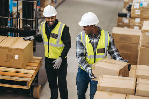 Glückliche Logistikmitarbeiter, die in einem Lagerhaus Kartons auf einen Palettenhubwagen laden. Zwei junge Männer lächeln fröhlich, während sie in einem großen Vertriebszentrum zusammenarbeiten. - JLPPF01414