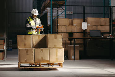Ein Lagerarbeiter liest ein Klemmbrett, während er hinter einem mit Kartons beladenen Palettenhubwagen steht. Ein Mann in einer reflektierenden Jacke nimmt Pakete zur Einlagerung in einem Vertriebszentrum entgegen. - JLPPF01415