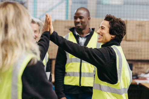 Cheerful warehouse workers high fiving each other during a staff meeting. Group of happy employees celebrating their success as a team in a distribution warehouse. - JLPPF01432
