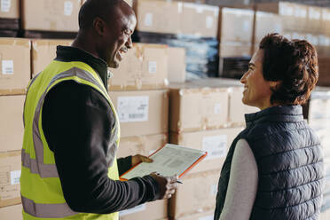 Happy logistics worker smiling at a female warehouse manager while holding a bill of lading. Cheerful young man delivering goods for shipment to a distribution centre. - JLPPF01436