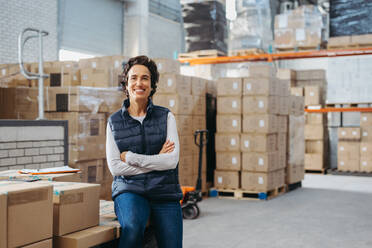 Mature warehouse supervisor smiling at the camera while sitting on a rack of cardboard boxes with her arms crossed. Happy woman working in a large distribution centre. - JLPPF01442