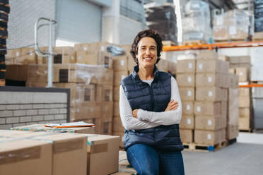 Mature warehouse employee smiling at the camera while sitting on a rack of package boxes with her arms crossed. Happy woman working as a manager in a large fulfillment centre. - JLPPF01443