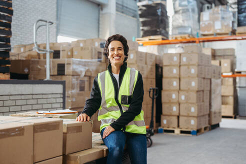 Cheerful warehouse supervisor smiling at the camera while sitting on a rack of package boxes in a reflective jacket. Happy mature woman working in a large fulfillment centre. - JLPPF01445