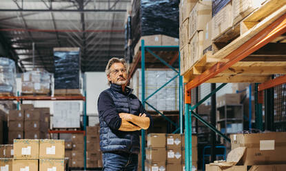 Älterer Lagerleiter, der mit verschränkten Armen dasteht und nachdenkt. Älterer Mann, der in einem großen Distributionszentrum über eine Lieferung nachdenkt. - JLPPF01446