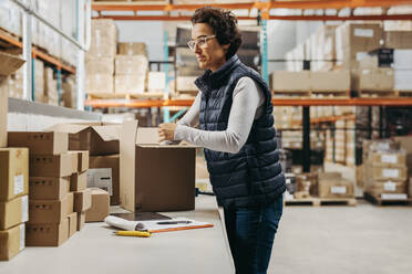 Mature warehouse worker packing a cardboard box in a distribution centre. Female logistics employee preparing an online order for shipment in a large fulfillment centre. - JLPPF01448