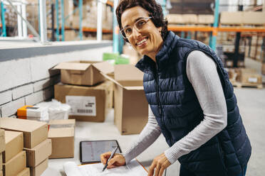 Happy warehouse manager writing a packing list while fulfilling orders in a logistics centre. Cheerful woman processing customer orders for shipment in a large distribution centre. - JLPPF01450