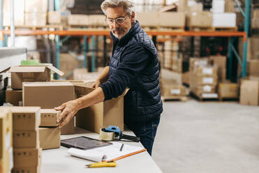 Ein älterer Mann benutzt ein digitales Tablet, während er in einem Auslieferungslager Aufträge verpackt. Ein älterer Logistikmitarbeiter benutzt eine Lagerverwaltungssoftware, um Kundenaufträge auszuführen. - JLPPF01457