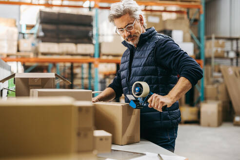 Senior man sticking scotch tape on a cardboard box in a distribution warehouse. Senior logistics worker preparing online orders for shipment in a large fulfillment centre. - JLPPF01458