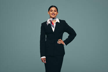 Selbstbewusste Stewardess, die in die Kamera lächelt, während sie in einem Studio steht und die Hand in die Hüfte stemmt. Freundliche Flugbegleiterin in Uniform vor einem Studiohintergrund. - JLPPF01474