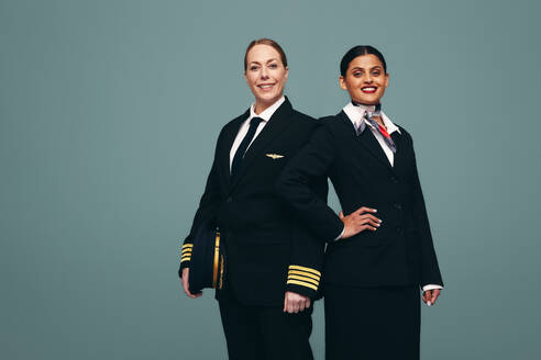 Glücklicher Flugkapitän und Stewardess lächeln in die Kamera, während sie zusammen stehen. Fröhlicher Pilot und Flugbegleiterin in ihren Uniformen vor einem Studiohintergrund. - JLPPF01504