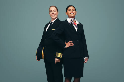 Selbstbewusster Pilot und Flugbegleiterin lächeln in die Kamera, während sie zusammen stehen. Fröhlicher Flugkapitän und Stewardess in ihren Uniformen vor einem Studiohintergrund. - JLPPF01505