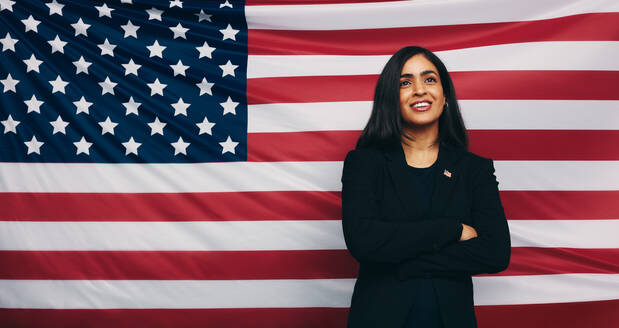 Nachdenkliche junge Kongressabgeordnete, die glücklich lächelnd vor der Flagge der Vereinigten Staaten von Amerika steht. Patriotische Politikerin, die mit verschränkten Armen steht. - JLPPF01512