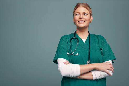 Medical doctor looking away thoughtfully while standing against a studio background. Happy young surgeon wearing green scrubs and a stethoscope around her neck. - JLPPF01533